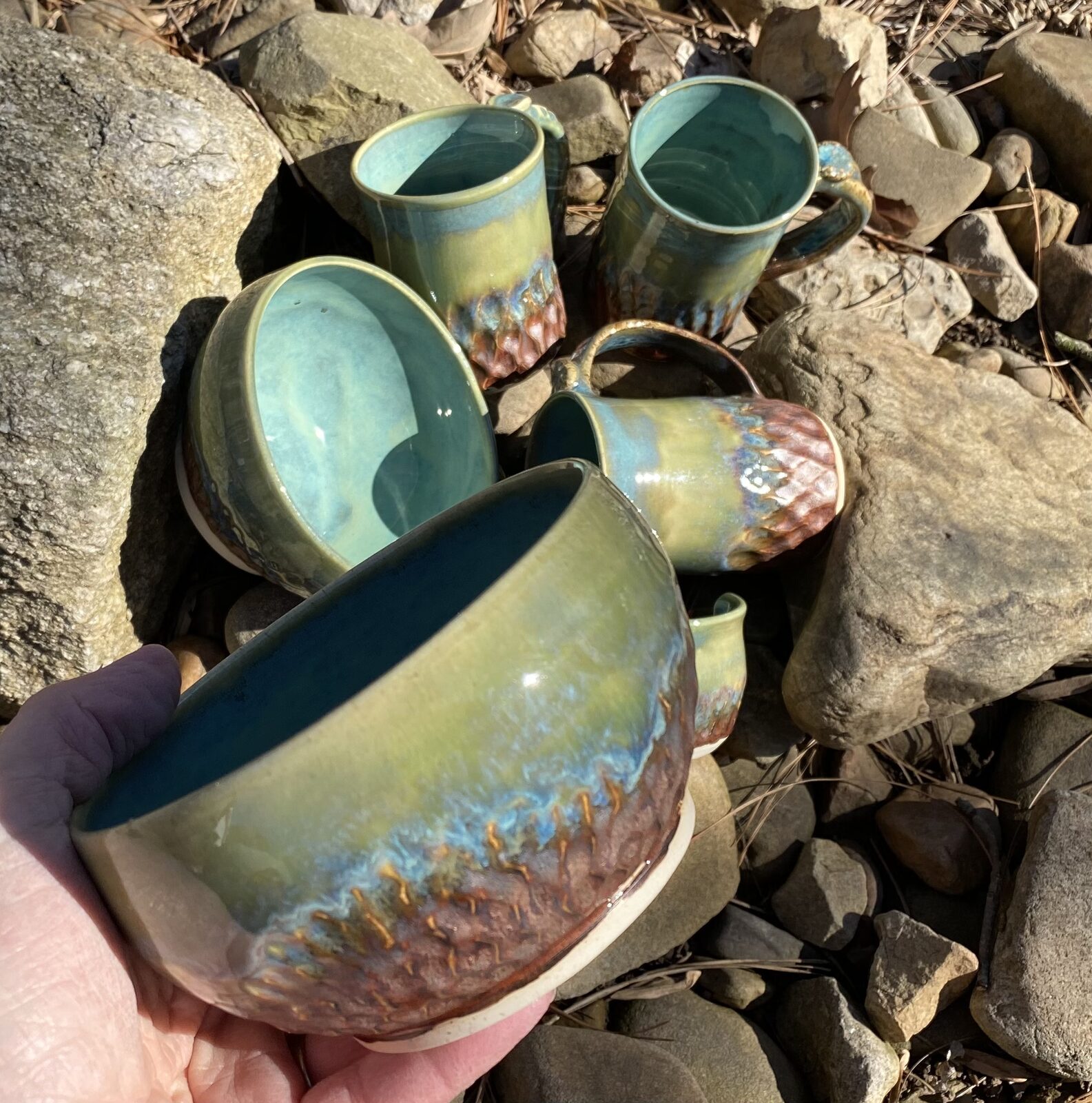 Set of mugs and bowls with matching green and copper drip glaze, photographed outdoors on rocks