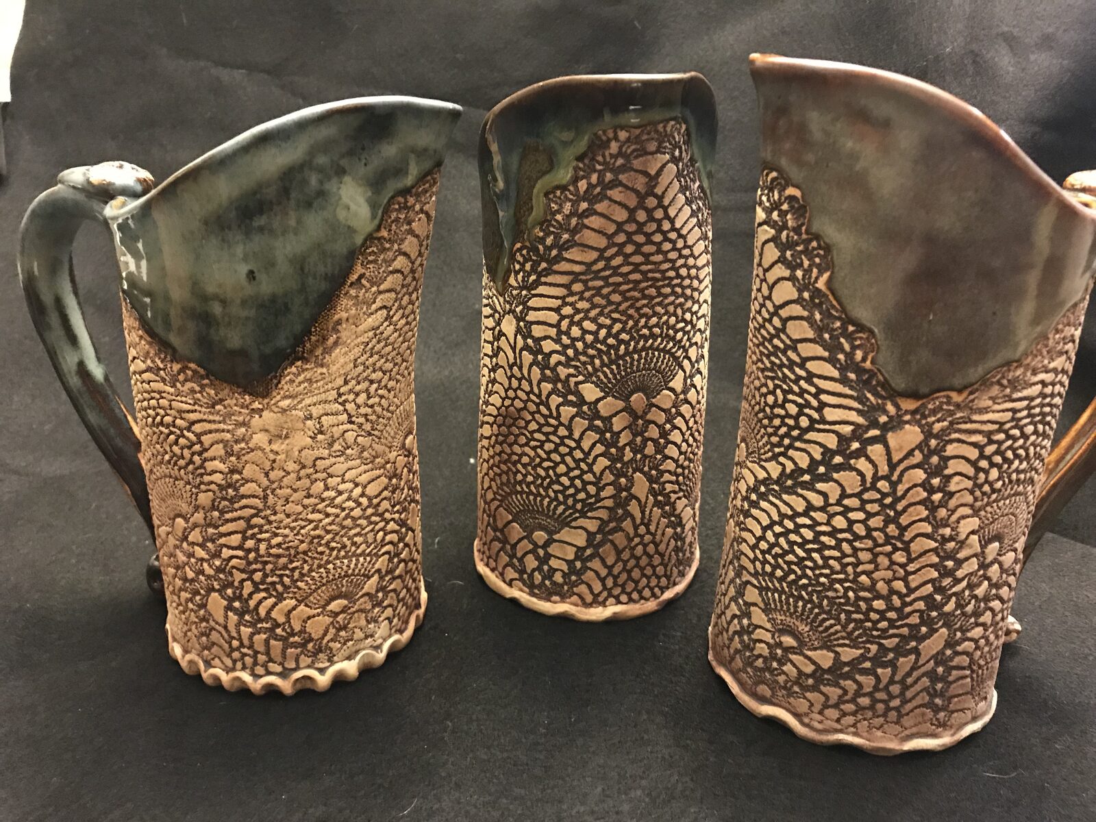 Three hand-built pitchers in a row with lace-textured raw clay bottoms and glazed tops
